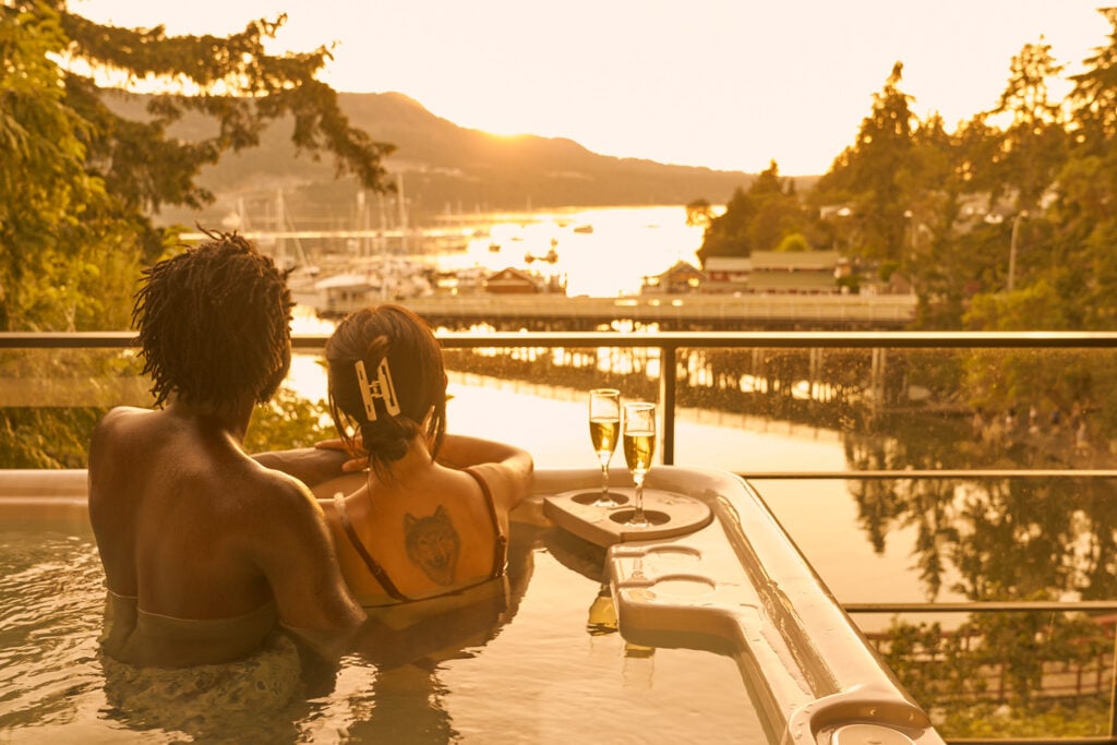 Couple relaxing in a private outdoor hot tub on a balcony, overlooking a marina during sunset
